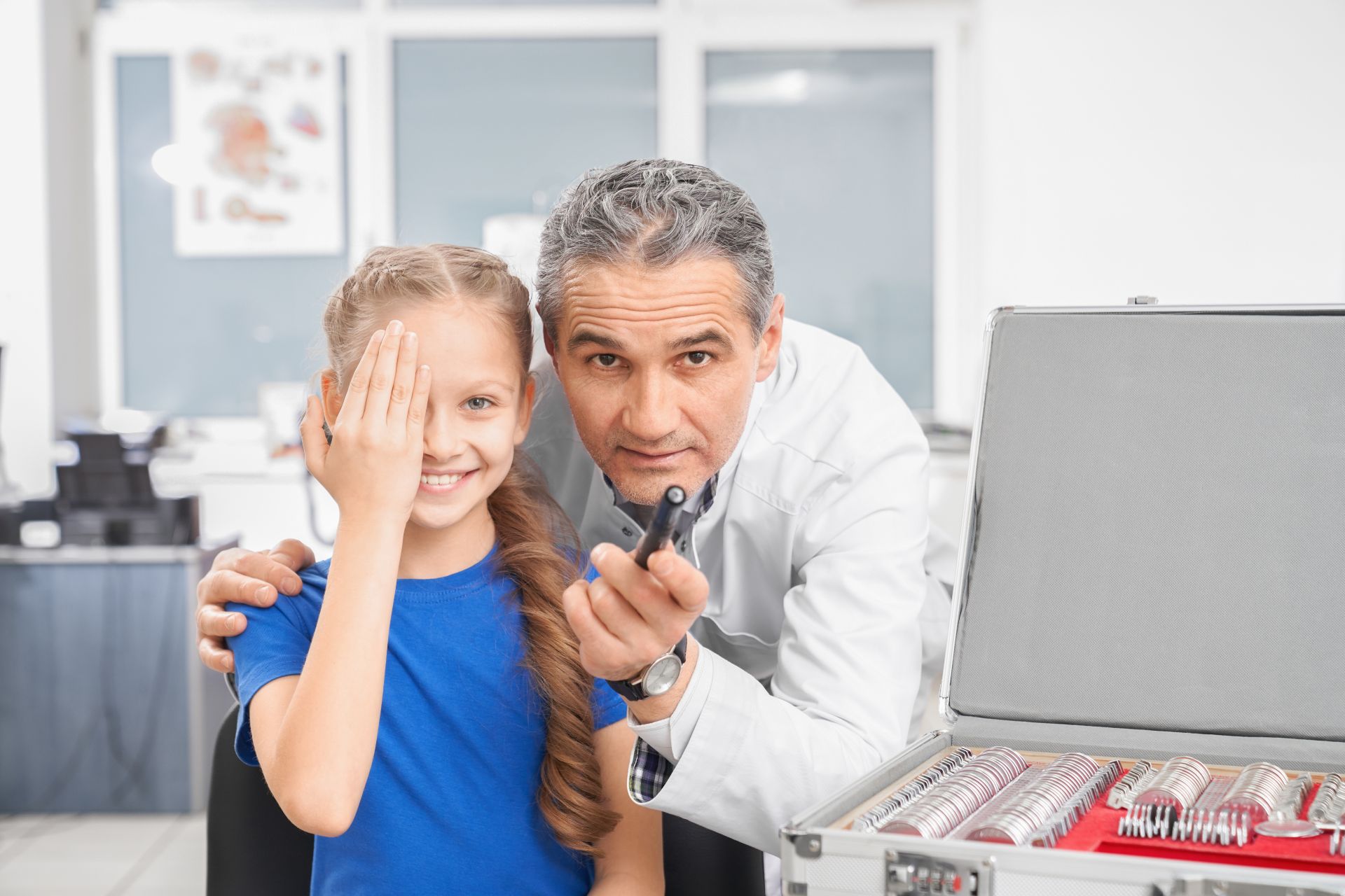 Front view of professional male oculist testing vision of little girl in clinic. Smiling girl closing eye with hands and looking at camera before choosing lens. Concept of eyewear and health.